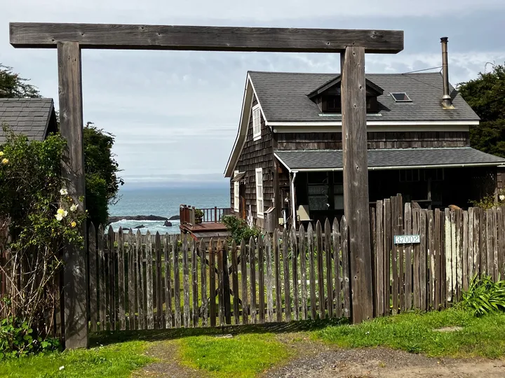 The weathered timber gate framing the shingled house and the Pacific beyond